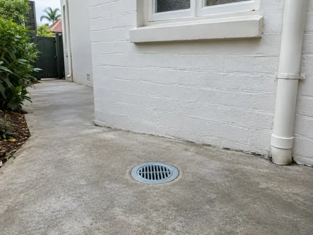 Concrete side path with a round grated drain beside a white brick wall in Helensburgh