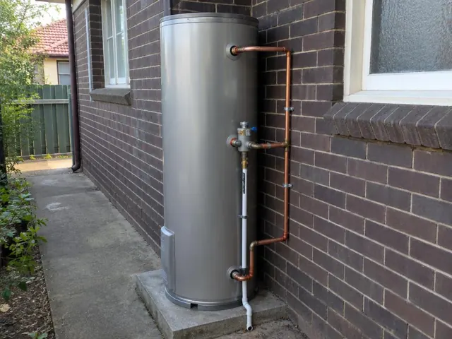 Outdoor hot water cylinder beside a brick wall with copper pipework and a drain line in Helensburgh